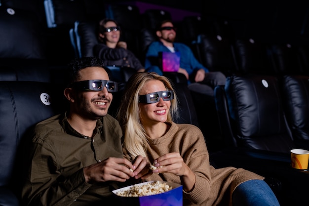 Cheerful couple in 3d eyeglasses eating popcorn while sitting in armchairs in front of large screen in dark cinema