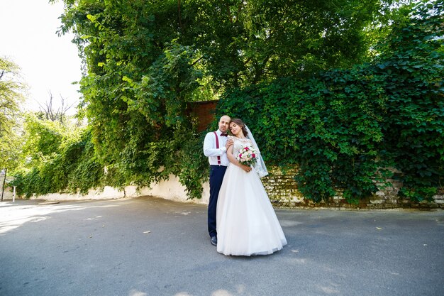 Sposa coppia allegra in un abito bianco con un bouquet mentre lo sposo con bretelle e papillon. sullo sfondo di un muro con foglie verdi. coppia felice. il concetto di matrimonio.