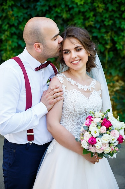 Cheerful couple bride in a white dress with a bouquet while the groom with suspenders and bow tie. Against the background of a wall with green leaves. Happy couple. The concept of marriage.