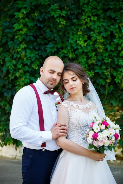 Cheerful couple bride in a white dress with a bouquet while the groom with suspenders and bow tie. Against the background of a wall with green leaves. Happy couple. The concept of marriage.