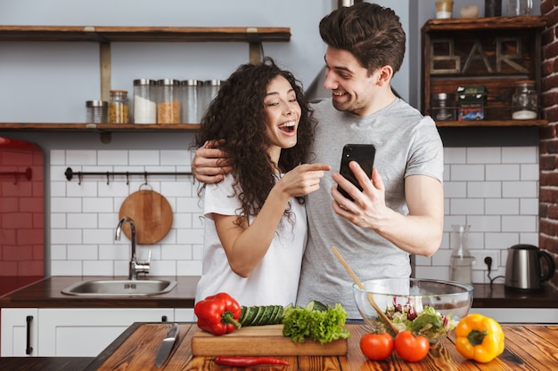 Coppia allegra che cucina una sana insalata fresca in cucina, si diverte, usa il cellulare