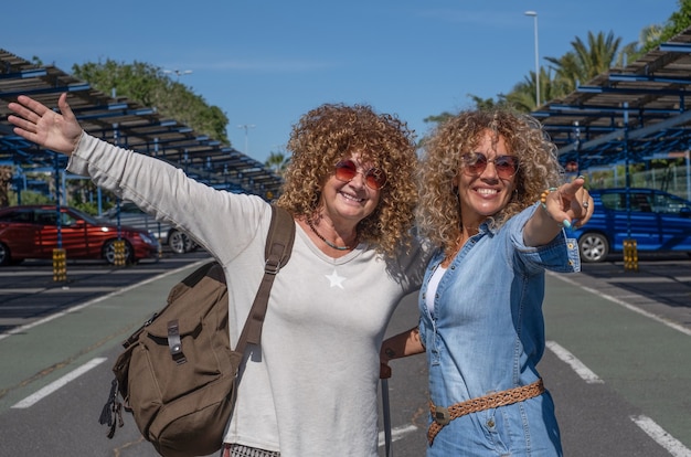 Cheerful couple of curly women in the parking of the airport with luggage and packpack ready to travel. smiling looking at camera