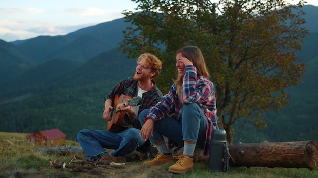 Cheerful couple enjoy nature at evening bonfire closeup Young travelers play guitar in summer mountains Smiling campers spend time together on vacation Family holiday relax leisure travel concept