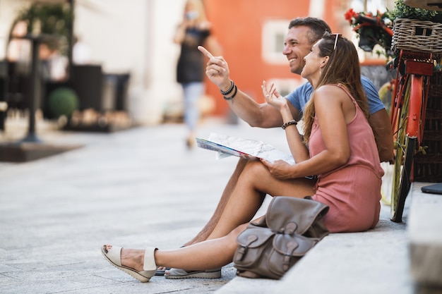 Coppie allegre che si divertono e si godono le vacanze estive seduti sulle scale ad esplorare la guida e la mappa di una città mediterranea.