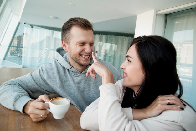 一杯のコーヒーと楽しい時間を過ごしている陽気なカップル。幸せな黒髪の女性は男の鼻に彼女の指で指さし、笑顔。カプチーノのカップを目を細めて保持している男性。