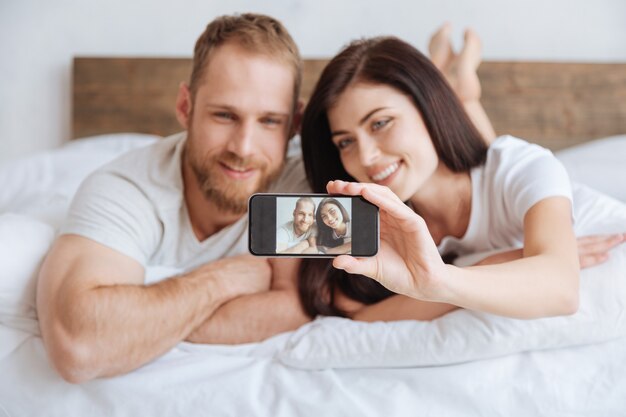 一緒にベッドに横たわって携帯電話で自分自身を撮影している陽気なカップル