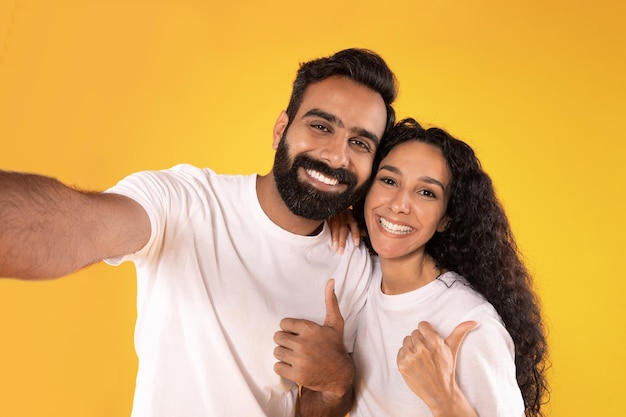 Cheerful couple making selfie gesturing thumbs up over yellow background