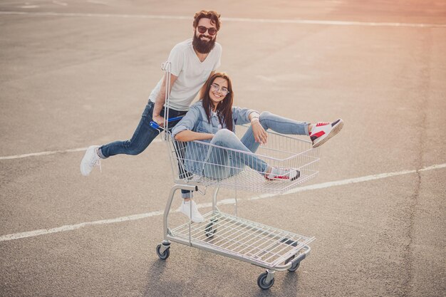 Cheerful couple riding shopping cart on parking