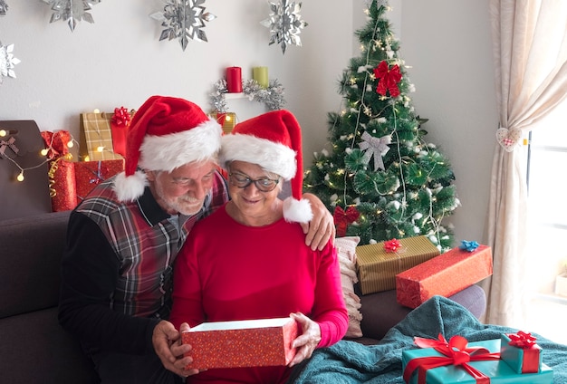 Allegra coppia di anziani seduti sul divano con cappelli di babbo natale che aprono un regalo con albero di natale e regali per la famiglia sullo sfondo. amore e concetto di famiglia