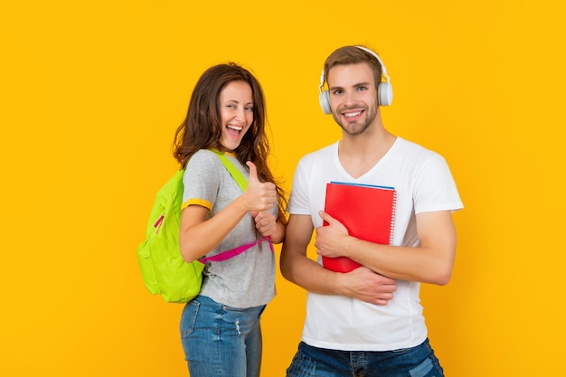 Cheerful couple of students in headphones with workbook and backpack on yellow background show thumb up, knowledge