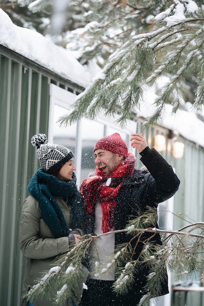 陽気なカップルが話し、近くの木々から雪が降っています。どちらも帽子、ジャケット、スカーフを着用しています。波形のシート壁のある家の前。