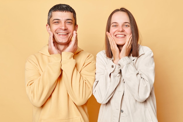 Cheerful couple woman and man wearing casual style clothing standing isolated over beige background smiling to camera keeping hands on cheeks