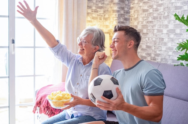 リビング ルームの快適なソファに座ってテレビでフットボールの試合を見ている若い男の子と年配の祖母のサッカー ファンの陽気なカップル