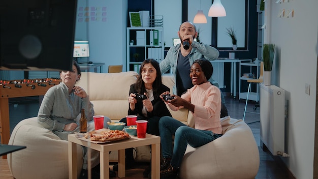 Cheerful coworkers playing video games with joysticks and\
console on television. workmates winning game with controllers on\
tv for entertainment, enjoying drinks and snacks after work