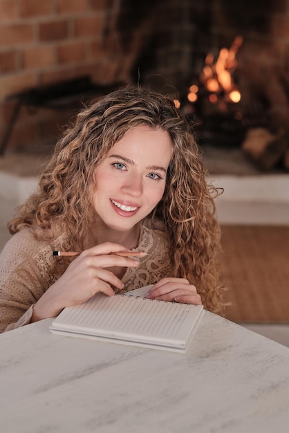 Cheerful creative female with pencil writing in diary while sitting in cozy living room near fireplace at home