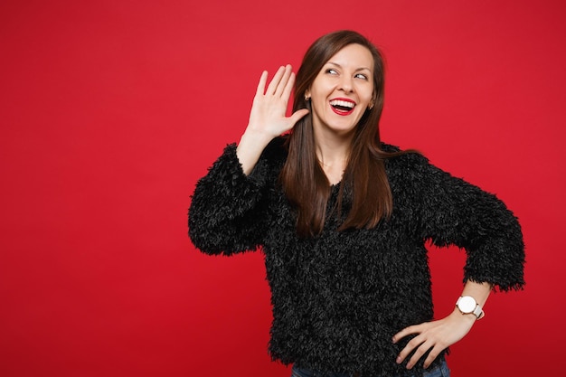 Cheerful curious young woman in black fur sweater eavesdrop with hearing gesture near ear isolated on bright red background in studio. People sincere emotions, lifestyle concept. Mock up copy space.