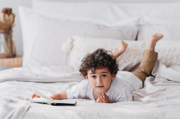 Cheerful curly american kid smiling laying on bed with book\
learning to read at home knowledge for preschoolers and education\
concept mockup pretty children everyday activities home\
education