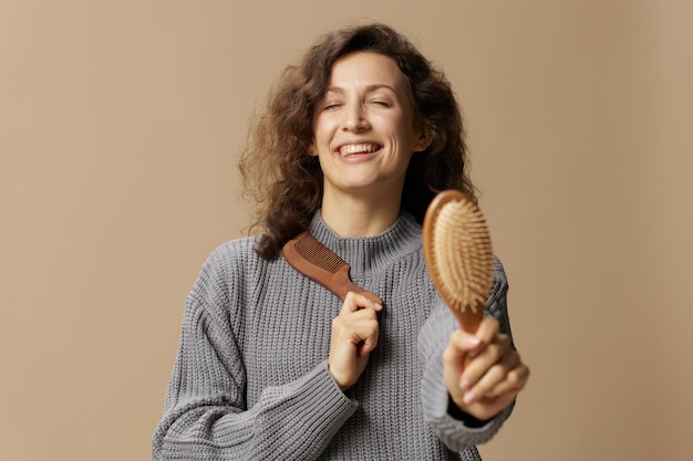 Cheerful curly beautiful female in gray casual sweater pulls hairbrush comb at camera recommended posing isolated on over beige pastel background Problematic unruly damaged hair concept Copy space