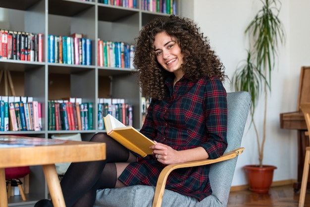 Ragazza castana riccia allegra che tiene un libro in sua mano mentre sedendosi nella biblioteca e nell'esaminare la macchina fotografica