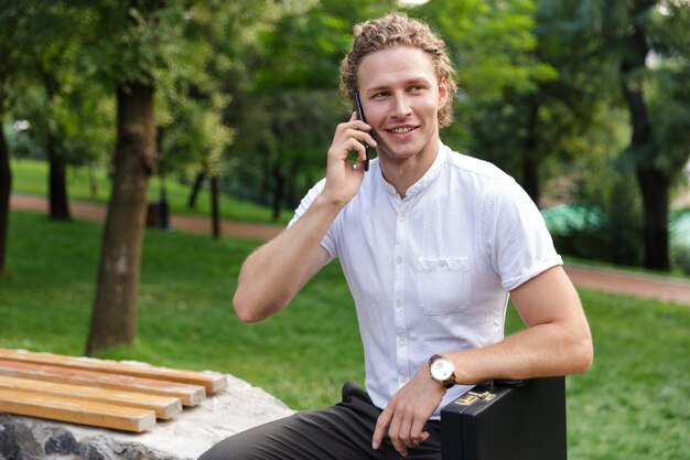 Cheerful curly business man with briefcase talking by smartphone and looking away while sitting on bench in park