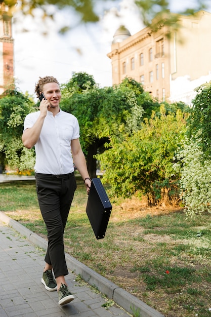 Cheerful curly business man with briefcase talking by smartphone while walking on the street