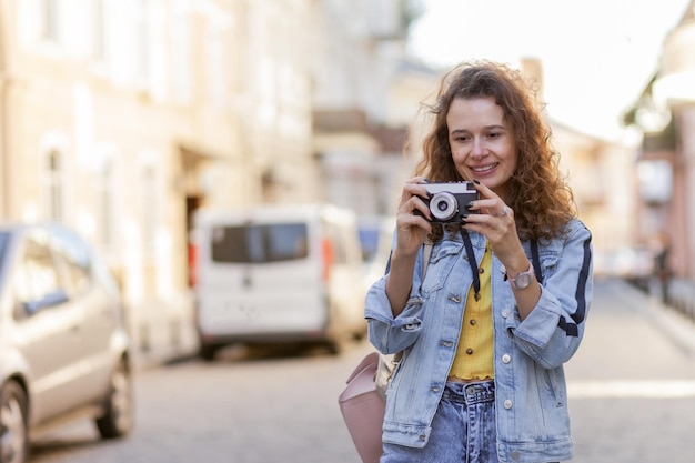 Cheerful curly hair woman tourist with retro camera walking along city street on bright sunny day Travel concept