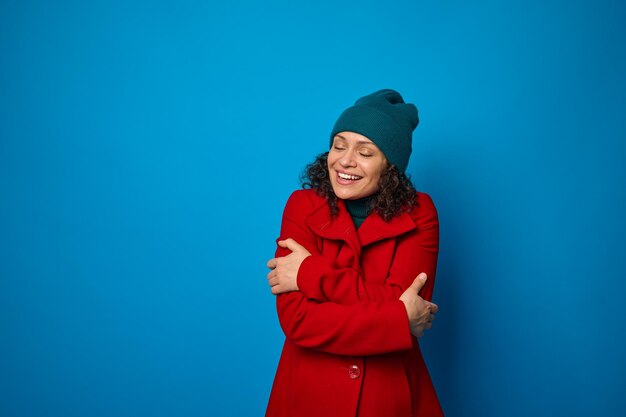 Cheerful curly-haired brunette pretty woman dressed in bright red coat and warm woolen green hat hugging warming herself poses with closed eyes and toothy smile against blue background with copy space