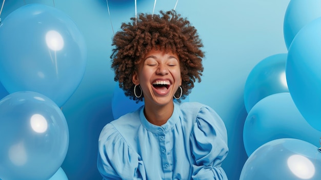 Веселая женщина с кудрявыми волосами, одетая в синюю одежду, смеется вокруг надутых гелийных воздушных шаров на заднем плане.