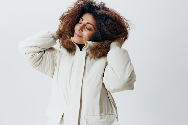 Cheerful curly pretty latin lady wearing fur hood of soft warm\
beige winter coat posing isolated closing eyes at white studio\
background fashion huge sale new collection concept horizontal side\
shot