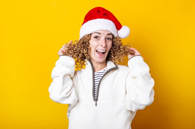 Cheerful curly young woman in santa claus hat on a yellow background