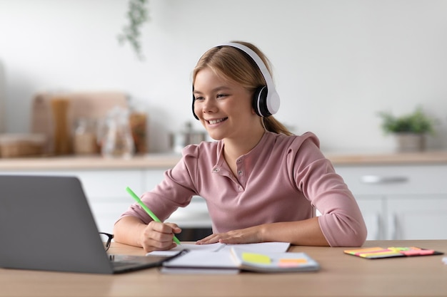 Cheerful cute caucasian teenager blonde girl student in headphones study at home watch video lesson