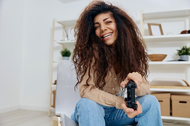Cheerful cute enjoyed tanned curly latin winner lady hold with\
joystick gamepad in both hands smile at camera sit on chair in home\
modern interior copy space mockup banner concept game platform