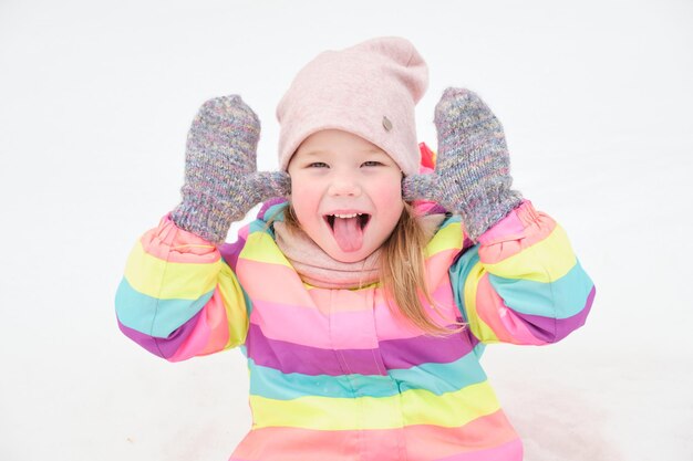 5 歳の陽気なかわいい女の子が雪の中で遊んで、面白い顔を作る