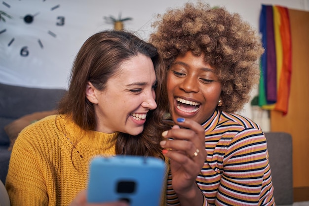 Photo cheerful cute lesbian couple laughing using cell phone in living room at home multiracial female