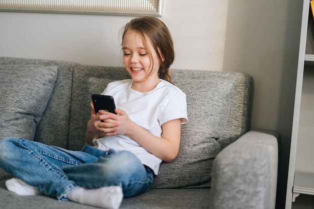 Cheerful cute little girl talking on video call by mobile phone in living room sitting on cozy sofa