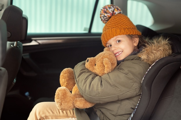Cheerful cute little girl in warm winterwear holding and embracing soft brown teddybear while sitting on backseat of car and looking at you