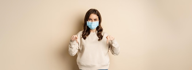 Cheerful cute woman in medical face mask pointing fingers down showing advertisement using protection from covid19 beige background