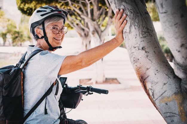 Cheerful cyclist elderly woman in urban park wearing helmet and backpack running with her electro bicycle Concept of healthy lifestyle and sustainable mobility