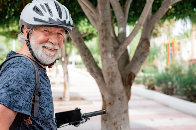 ヘルメットと電気自転車で走るバックパックを身に着けている都市公園の陽気なサイクリストの年配の男性健康的なライフ スタイルと持続可能なモビリティの概念
