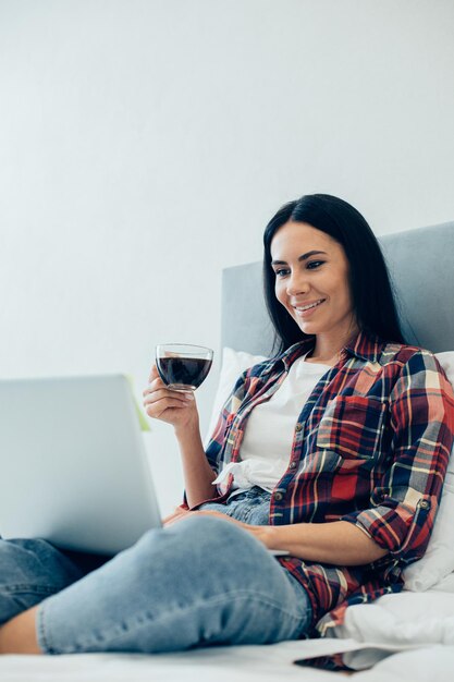 Cheerful dark haired woman enjoying hot coffee from a glass cup and smiling while looking at the laptop screen