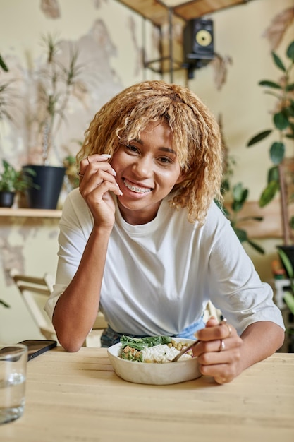 Photo cheerful dark skinned girl with braces holding fork near fresh salad bowl in modern vegan cafe