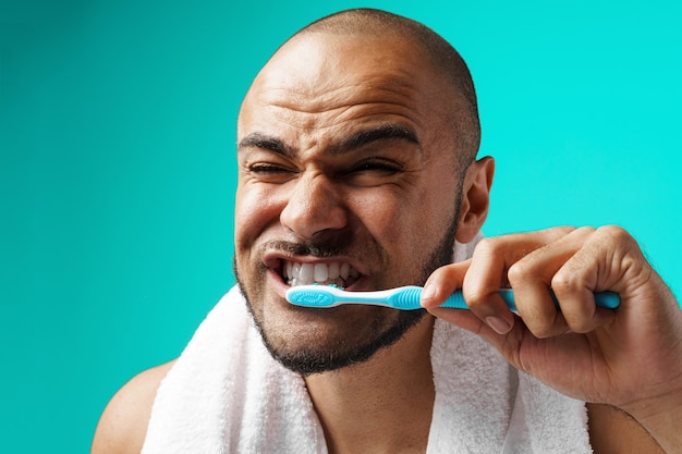 Cheerful dark-skinned male brushing his teeth on turquoise