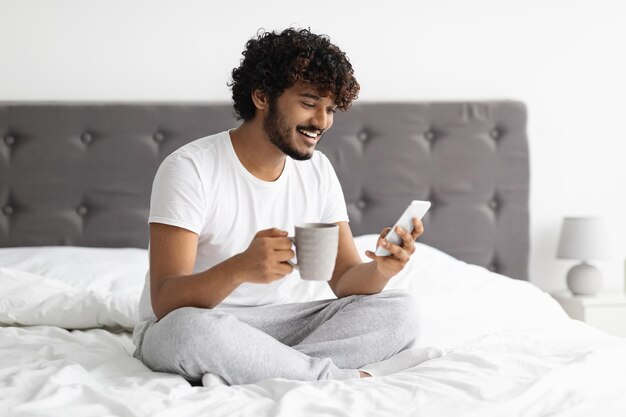 Cheerful darkskinned man drinking coffee and using smartphone