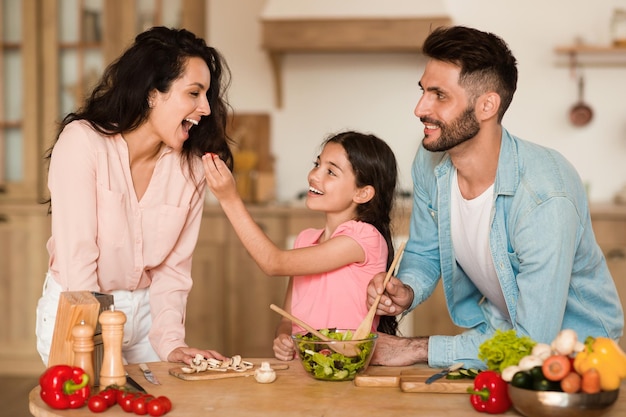 Cheerful daughter feeding her happy mother with small cherry tomato while preparing salad together