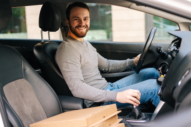 Foto il fattorino allegro che indossa abiti casual alla guida dell'auto ha consegnato la pizza calda al cliente, guardando la telecamera. concetto di consegna online veloce in giro per la città.