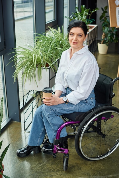 Foto allegra donna d'affari disabile in abbigliamento elegante in sedia a rotelle con il caffè in mano e guardando la telecamera