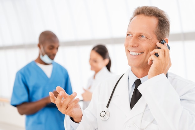 Cheerful doctor on the phone. Cheerful mature doctor talking on the mobile phone and gesturing while his colleagues talking in the background