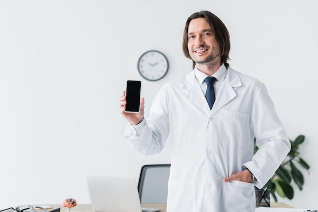 Photo cheerful doctor in white coat looking at camera and holding smartphone in hand