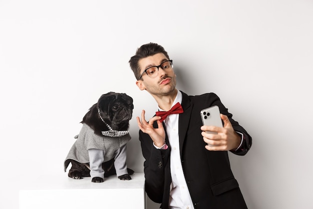 Cheerful dog owner in suit celebrating New Year with dog, taking selfie on smartphone near cute black pug in costume, white background