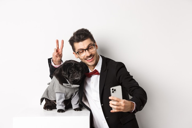 Cheerful dog owner in suit celebrating New Year with dog, taking selfie on smartphone near cute black pug in costume, white background